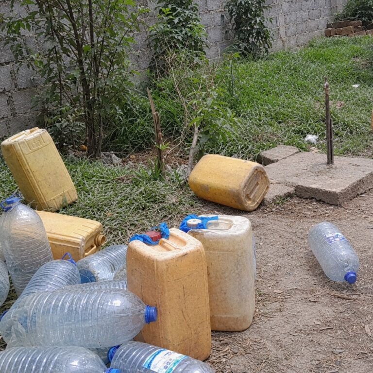 Bidons jaunes et bouteilles en plastique vides posés près d'un robinet sur dalle de béton, dans une cour herbeuse